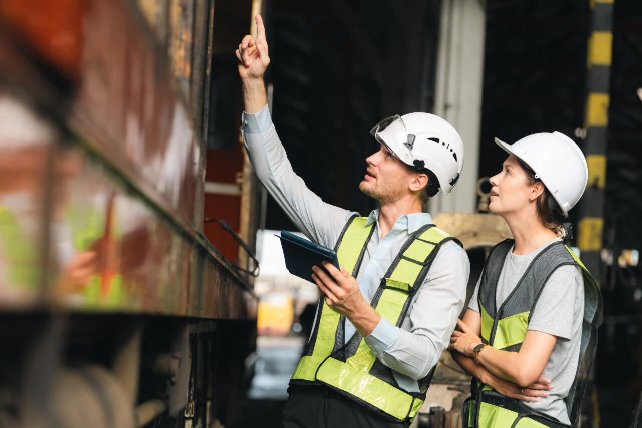 Engineers wearing safety helmets inspecting railway blueprint beside a vintage locomotive, symbolizing teamwork, innovation, and development in modern rail transportation and infrastructure projects