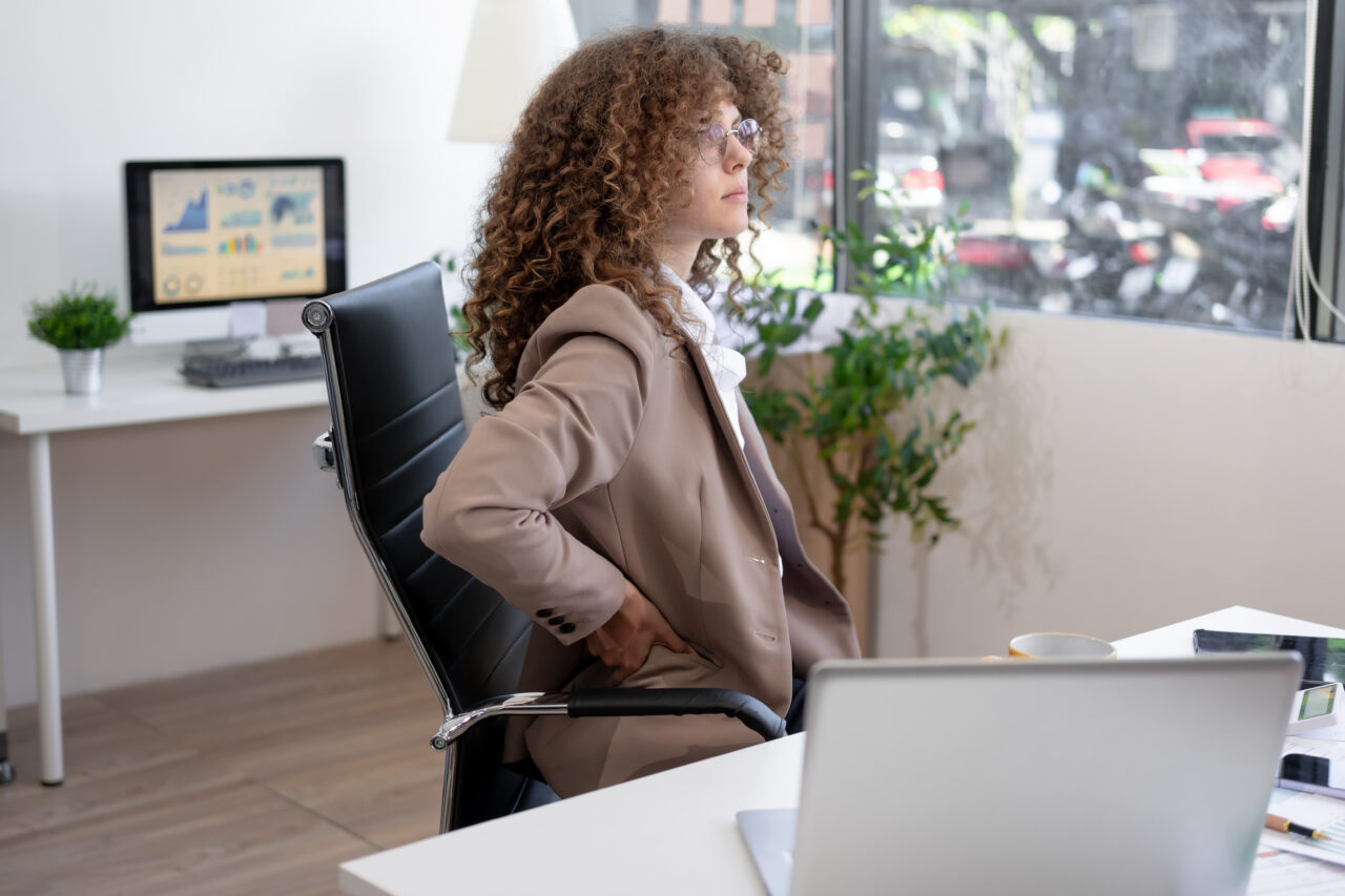 Exhausted professional woman stretching neck while working at office desk with laptop and paperwork, highlighting workplace fatigue, stress management, ergonomics, health awareness, productivity
